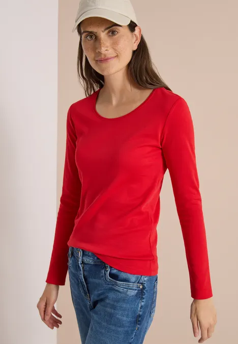 Basic shirt met lange mouwen en ronde hals flame red Eine Frau mit rotem Langarmshirt, blauen Jeans und beige Kappe vor neutralem Hintergrund lächelt sanft.