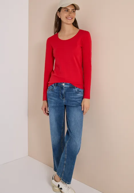Basic shirt met lange mouwen en ronde hals flame red Frau mit rotem Langarmshirt, blauen Jeans, weißen Sneakern und Basecap steht lächelnd in heller Ecke.