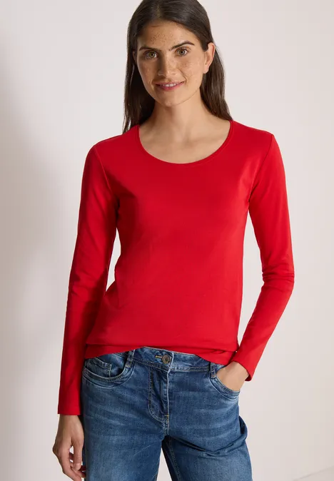Basic shirt met lange mouwen en ronde hals flame red Frau trägt rotes Langarmshirt und Jeans, steht mit Hand in Tasche und lächelt.