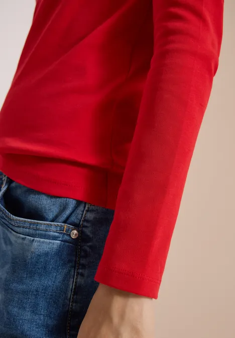 Basic shirt met lange mouwen en ronde hals flame red Nahaufnahme einer Person in rotem Langarmshirt und blauen Jeans, Fokus auf Arm und Taille