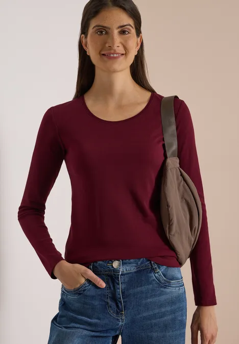 Basic Langarm Shirt mit Rundhals red wine Frau in dunkelrotem Langarmshirt, blauen Jeans und Schultertasche, die lächelnd vor beiger Wand steht