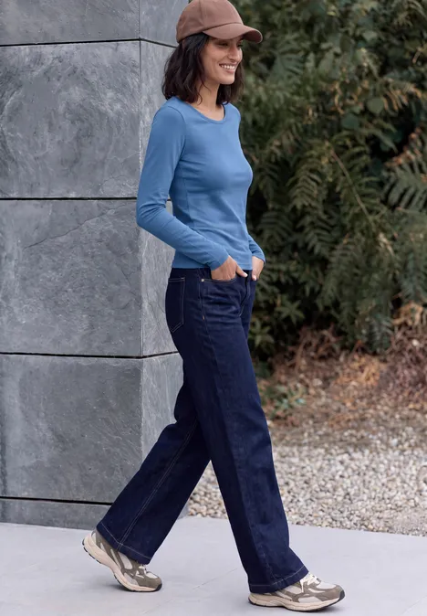 T-shirt basique à manches longues et col rond north blue Frau in blauem Langarmshirt, weiten Jeans und Cap geht draußen an einer Wand entlang.