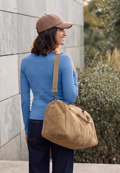 T-shirt basique à manches longues et col rond north blue Frau in blauem Oberteil, brauner Kappe und Jeans trägt große beige Tasche und steht vor einer Wand.