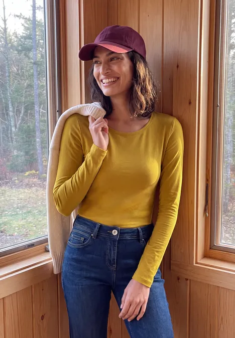T-shirt basique à manches longues et col rond golden honey Eine lächelnde Frau im gelben Oberteil, Jeans und roter Mütze hält eine Jacke in einer Holzhütte.