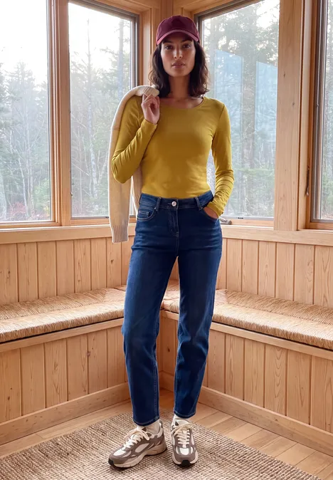 T-shirt basique à manches longues et col rond golden honey Frau mit gelbem Pullover, blauen Jeans und Mütze steht in einem hellen, holzvertäfelten Raum.