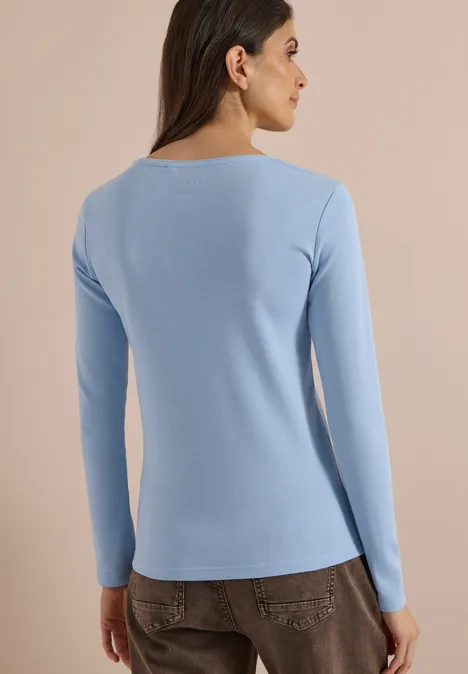 Frau von hinten in schlichtem hellblauen, langärmligen Shirt vor neutralem Hintergrund.