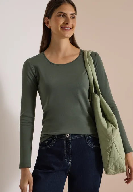 Lächelnde Frau in grünem Langarmshirt und gesteppter Schultertasche vor neutralem Hintergrund.