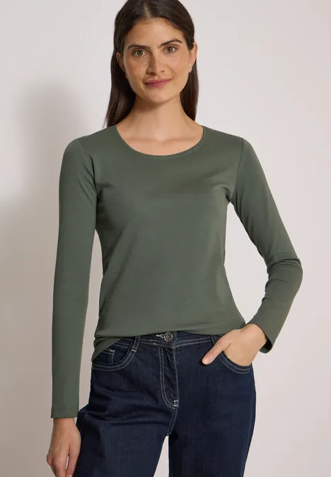 Junge Frau in schlichtem, olivgrünen Langarmshirt und blauen Jeans vor neutralem Hintergrund.
