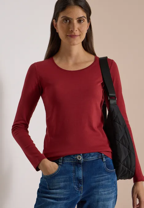Basic Langarm Shirt mit Rundhals russet red Eine Frau in rotem Langarmshirt, Jeans und schwarzer Umhängetasche vor hellem Hintergrund.