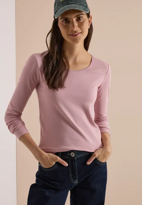 Femme souriante portant une casquette bleue, un t-shirt rose et un jean, mains dans les poches.