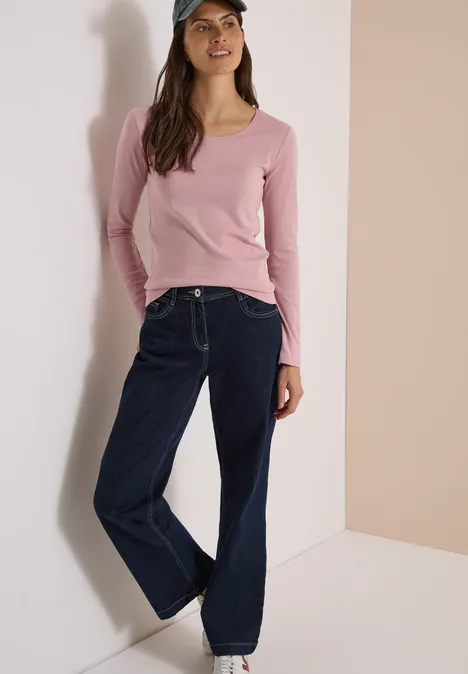 Une femme appuyée contre un mur porte un top rose, un jean large et une casquette.