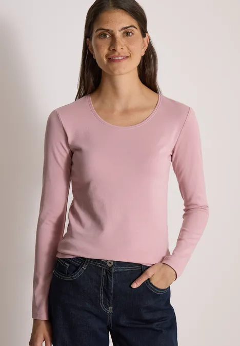 Une femme portant un haut à manches longues rose et un jean devant un mur clair.