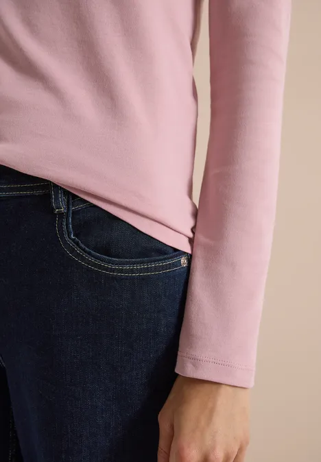 Gros plan sur une personne en haut rose manches longues et jean sombre, main le long du corps.
