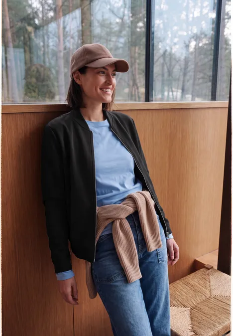 Une femme souriante en casquette, veste bleue et jean, appuyée sur une fenêtre dans un intérieur lumineux.