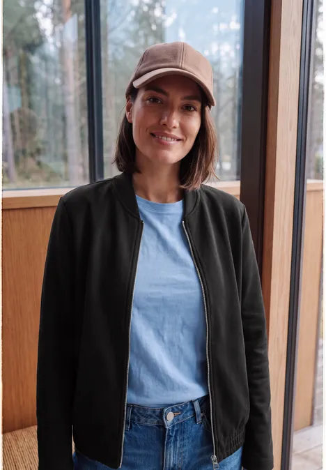 Jeune femme souriante en casquette, veste noire et jean, devant une grande fenêtre en bois.