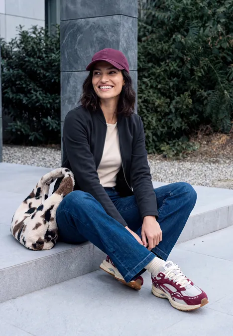 Jersey blouson met ritssluiting urban dark blue Jonge vrouw zit buiten op de trap, draagt pet, jas, jeans en sneakers en lacht.