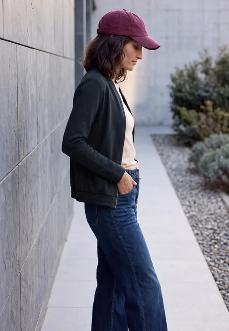Jersey blouson met ritssluiting urban dark blue Vrouw met pet, zwarte jas en spijkerbroek leunt zijwaarts tegen een buitenmuur.