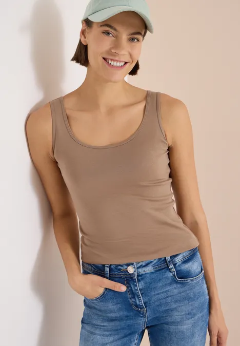 Jeune femme en débardeur beige, jean et casquette, pose décontractée devant un fond clair.