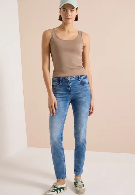 Femme en débardeur beige, jean bleu et baskets à motifs devant un fond neutre.