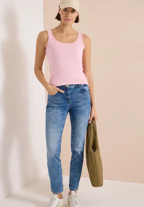Frau in pinkem Tanktop und blauen Jeans steht vor hellem Hintergrund und hält eine olivgrüne Jacke.