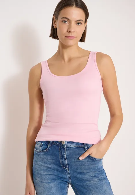 Frau trägt rosa Tanktop und blaue Jeans vor neutralem Hintergrund.