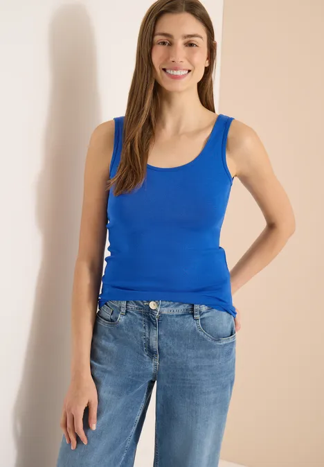 Frau in blauem Tanktop und Jeans vor neutralem Hintergrund.