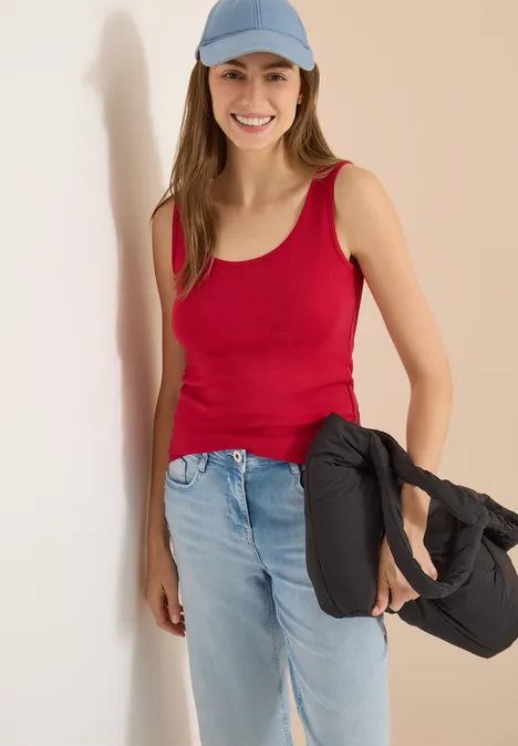 Frau mit rotem Top und Jeans lehnt an der Wand, trägt eine schwarze Tasche über der Schulter.