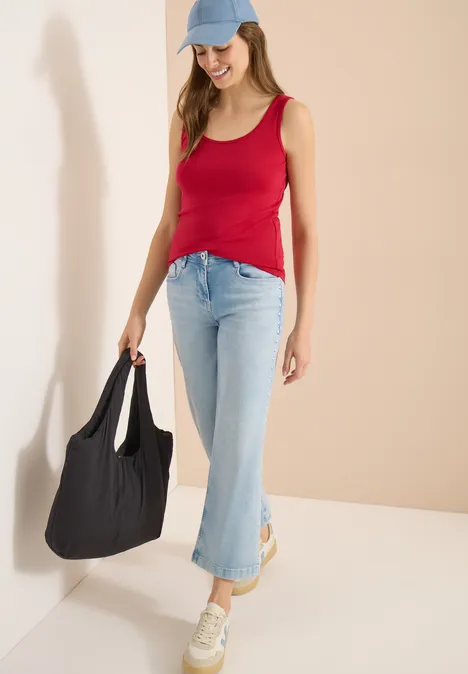 Frau in rotem Top, hellblauer Jeans, Sneakern und Cap, mit großer schwarzer Tasche.