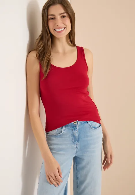 Eine Frau lehnt lässig an einer Wand und trägt ein rotes Tanktop und helle Jeans.