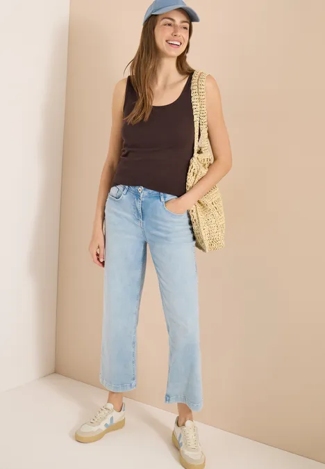 Frau trägt schwarzes Top, helle Jeans, Sneaker und Korbtasche vor neutralem Hintergrund.