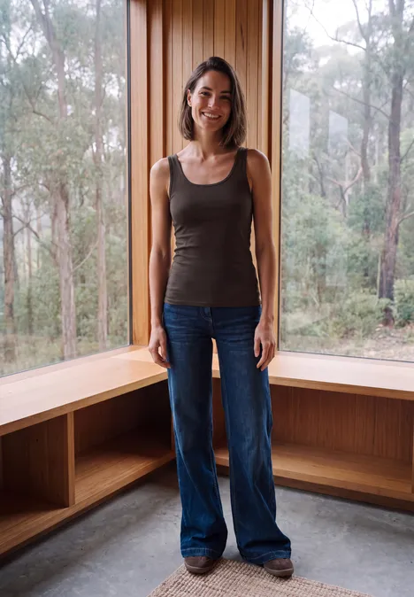 Basic top van puur katoen truffle brown Een vrouw in jeans en tanktop staat in een lichte houten ruimte voor grote ramen met uitzicht op bos.