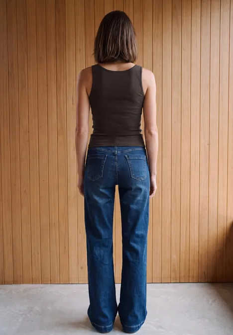 Basic top van puur katoen truffle brown Een vrouw van achteren, staand voor een houten wand, met een zwarte top zonder mouwen en een blauwe jeans.