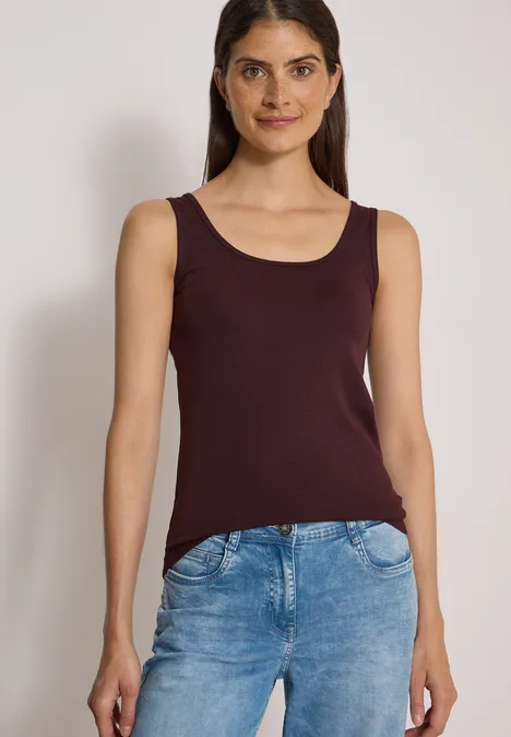 Basic top van puur katoen berry red Frau steht vor neutralem Hintergrund, trägt ein bordeauxfarbenes Top und blaue Jeans.