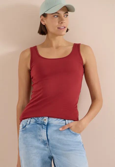 Frau mit rotem Top und blauer Jeans steht vor beigem Hintergrund, eine Hand in der Tasche.