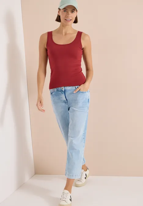 Frau in rotem Top, blauen Jeans und weißen Sneakern vor neutralem Hintergrund.
