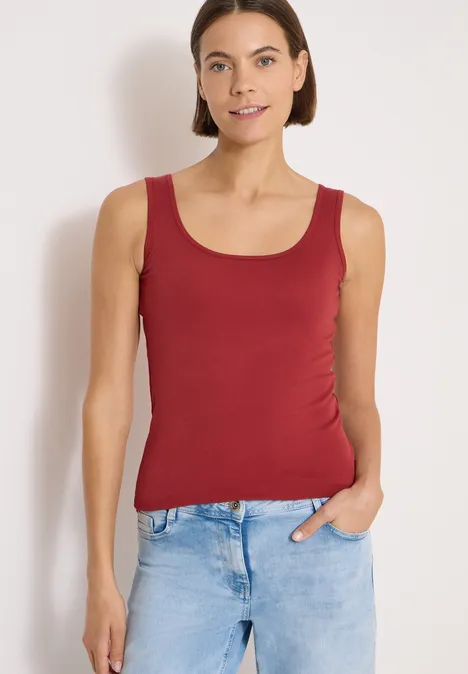 Frau in rotem ärmellosem Top und blauen Jeans vor hellem Hintergrund, eine Hand in der Tasche.