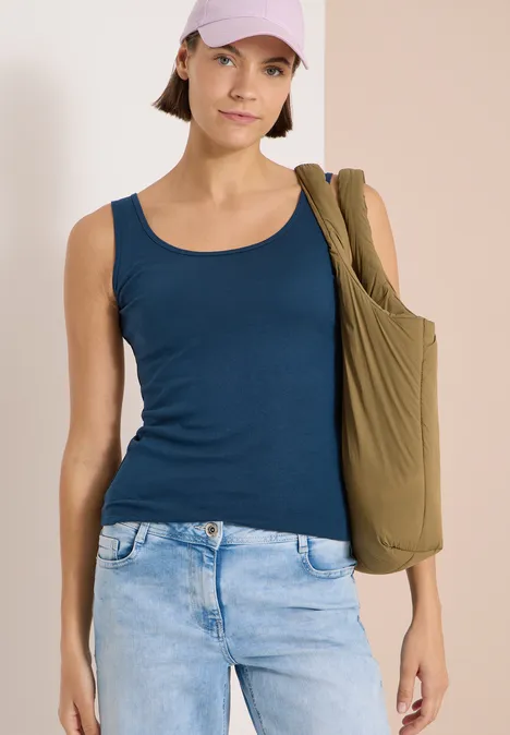 Frau in blauem Tanktop und hellblauen Jeans mit olivgrünem Schulterbeutel vor neutralem Hintergrund.