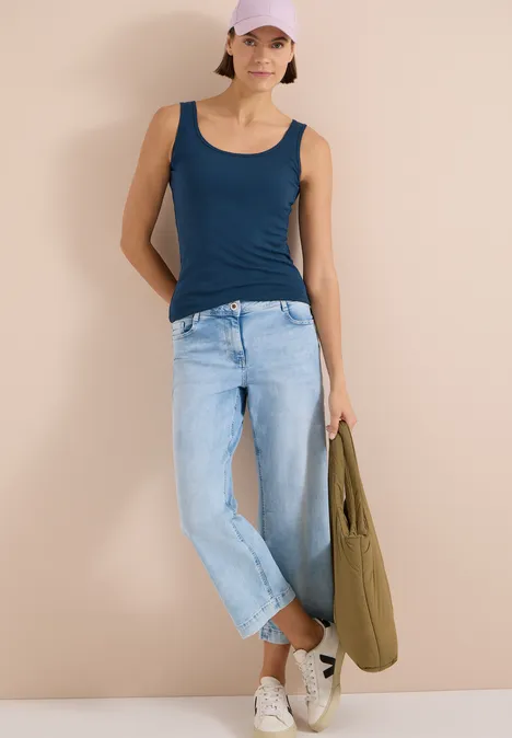 Eine Frau in blauem Top und heller Jeans lehnt an einer Wand und hält eine grüne Tasche.