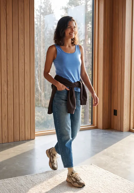 Basic Top aus reiner Baumwolle north blue Frau in blauem Top und Jeans steht in modernem Raum mit großen Fenstern.