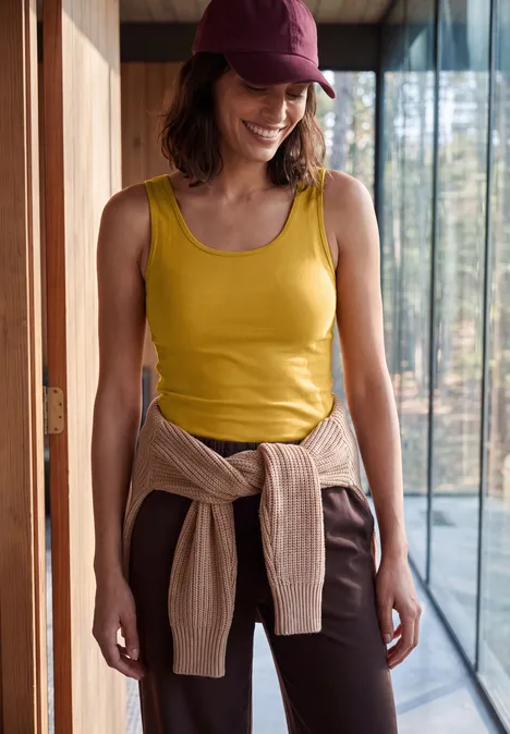 Frau in gelbem Tanktop, braunen Hosen und Cap steht lächelnd in modernem Haus, Pullover um die Taille.