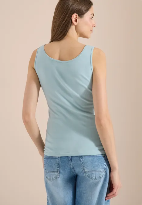 Femme vue de dos portant un débardeur bleu clair et un jean sur fond beige.