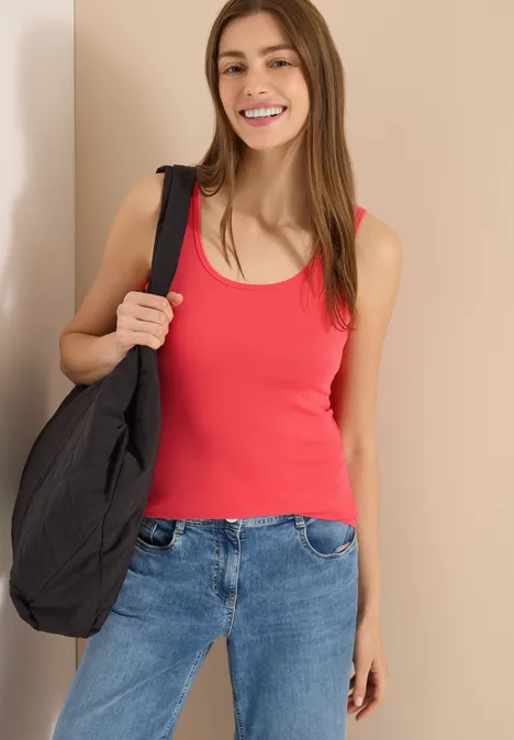 Frau in rotem Top, Jeans und schwarzer Umhängetasche vor hellem Hintergrund.