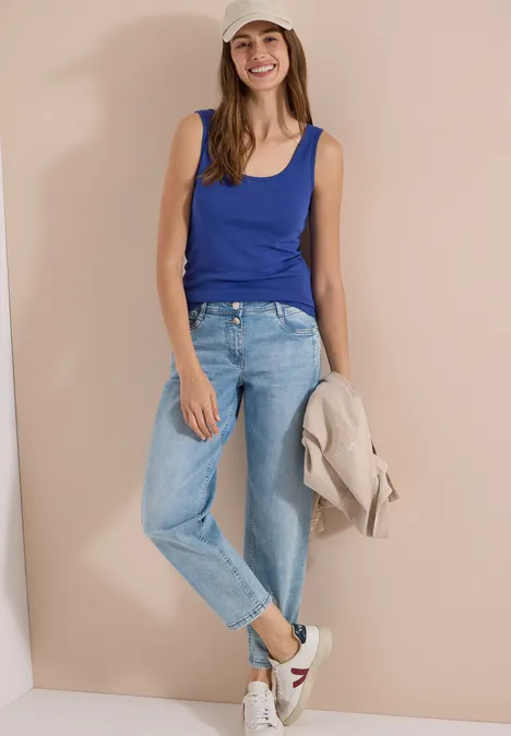 Junge Frau lehnt in blauem Top und Jeans an einer hellen Wand, hält beige Jacke und trägt weiße Sneaker.