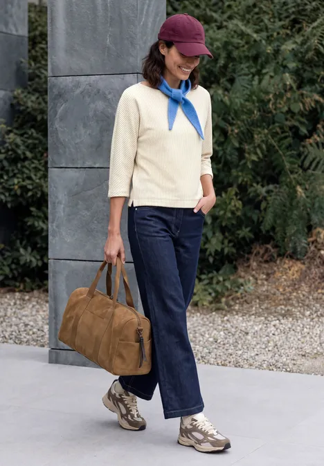 Frau in Jeans, cremefarbenem Pullover, blauem Halstuch und Cap, mit brauner Tasche spaziert vor grauer Fassade.