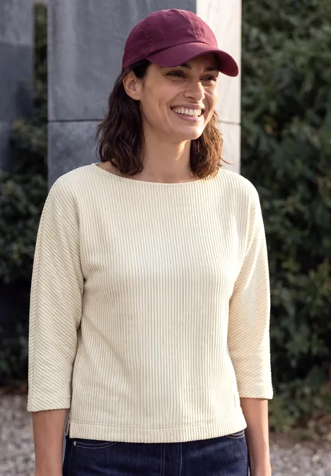 Lächelnde Frau mit rotem Cap und hellem Pullover vor Säulen und grünem Hintergrund.