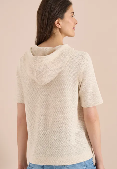 Frau trägt von hinten gesehen einen cremefarbenen, kurzärmeligen Strick-Hoodie vor neutralem Hintergrund.