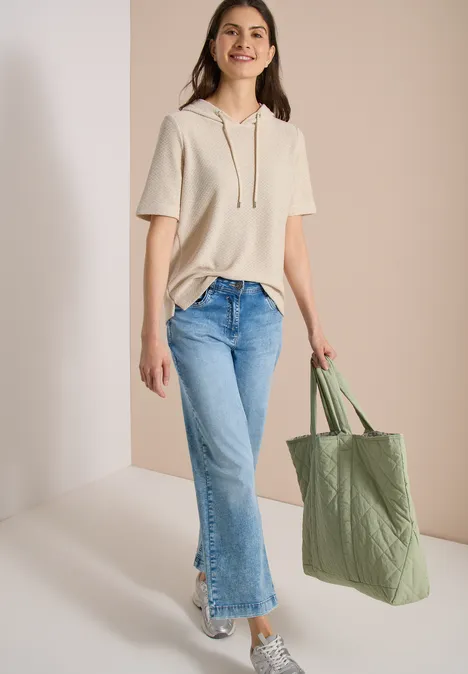 Frau in kurzem beigen Hoodie, hellen Jeans und Sneakers mit grüner Stofftasche.