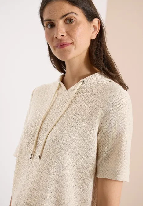 Frau trägt cremefarbenen, kurzärmeligen Strick-Hoodie mit Kordelzug.