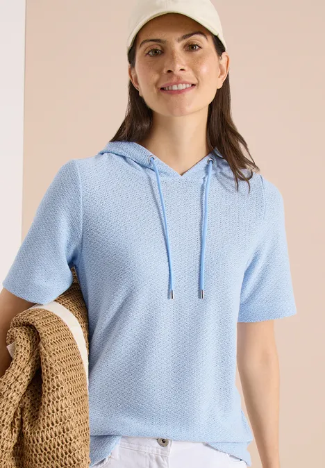 Frau trägt hellblaues Kurzarm-Kapuzen-Shirt in Strukturstrick, hält beigefarbenen gehäkelten Shopper.