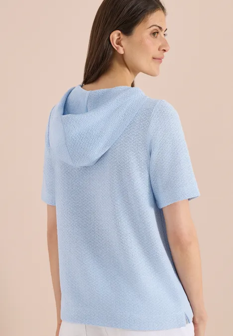 Rückansicht einer Frau in hellblauem, kurzärmeligem Strick-Hoodie vor hellem Hintergrund.